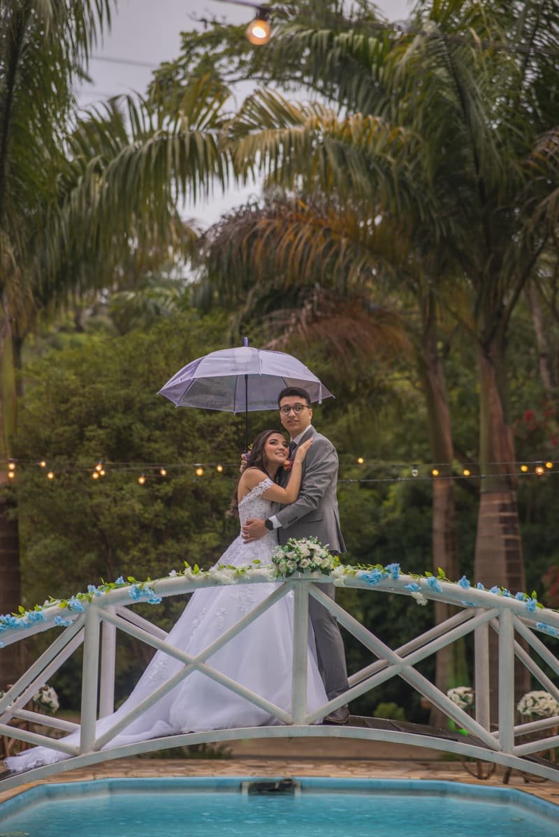 Casal em ensaio pré-wedding com guarda-chuva em jardim com piscina